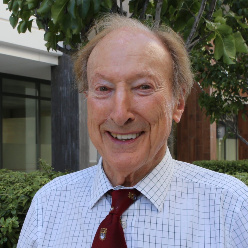 Man in blue checkered shirt and maroon tie smiling at camera outdoors with ivy background