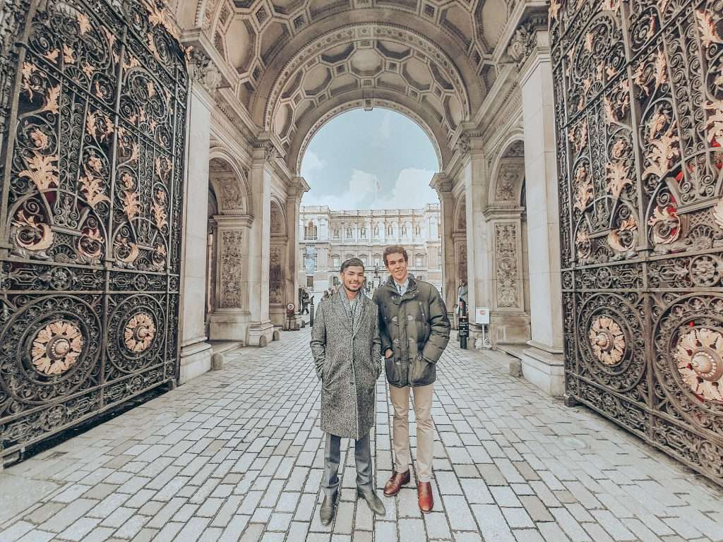 Two young men smiling at camera in ornate stone archway with decorative iron gates