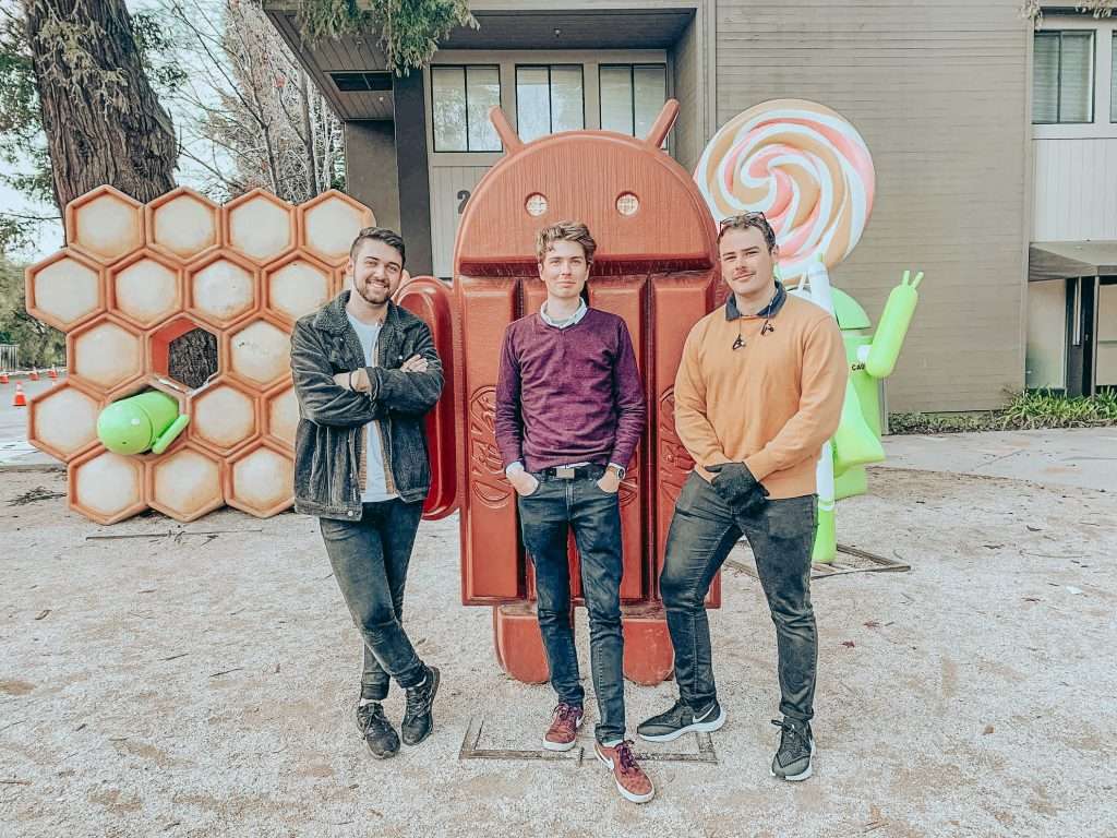 Three men posing in front of large red Android mascot statue and colorful geometric sculptures