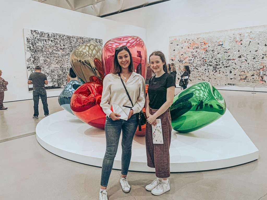 Two young women smiling at camera in front of red and green metallic sculptural art installations at a modern art gallery.