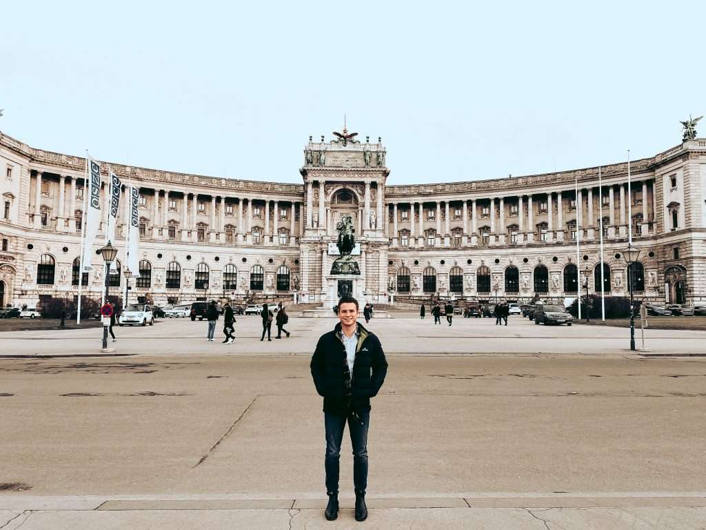 Man in black jacket standing in plaza in front of large ornate palace building in Vienna