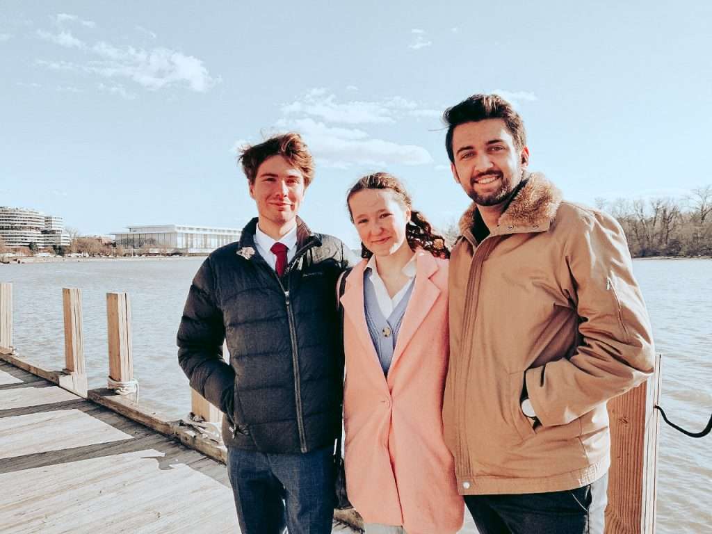 Three young adults standing on a dock by a river, smiling at camera with city buildings in background