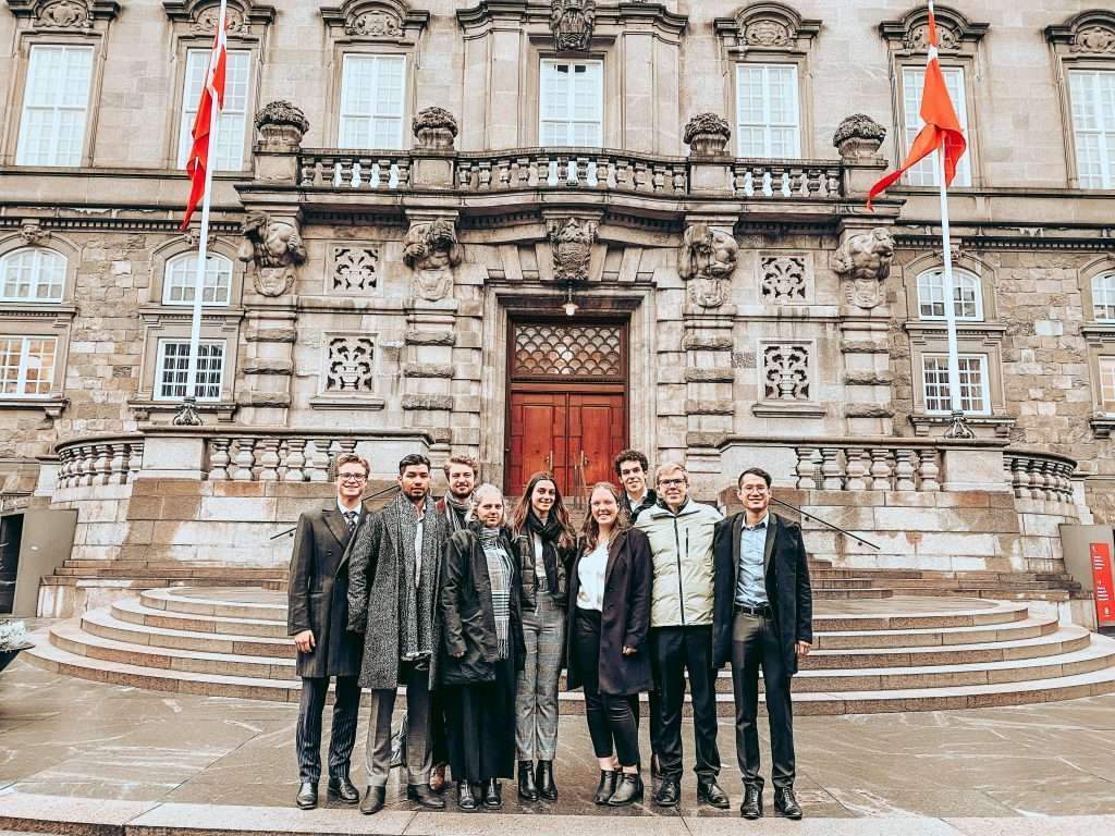 Nine professionals standing together on steps in front of a historic stone building with red flags and classical architecture.