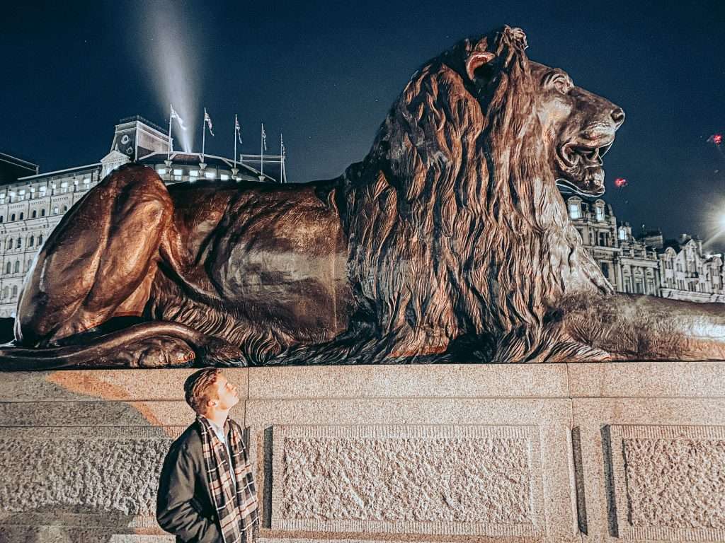Young man looking up at large bronze lion statue at night with city buildings behind