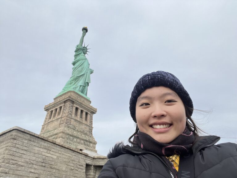 Young woman smiling at camera with Statue of Liberty in background, wearing winter coat and black beanie