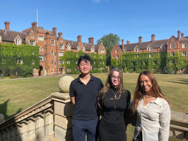 Three young adults smiling at camera in front of historic red brick college buildings covered in ivy