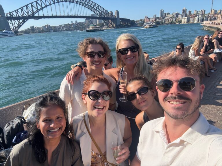 Group of six smiling people posing with champagne by Sydney Harbour with the Sydney Harbour Bridge in background