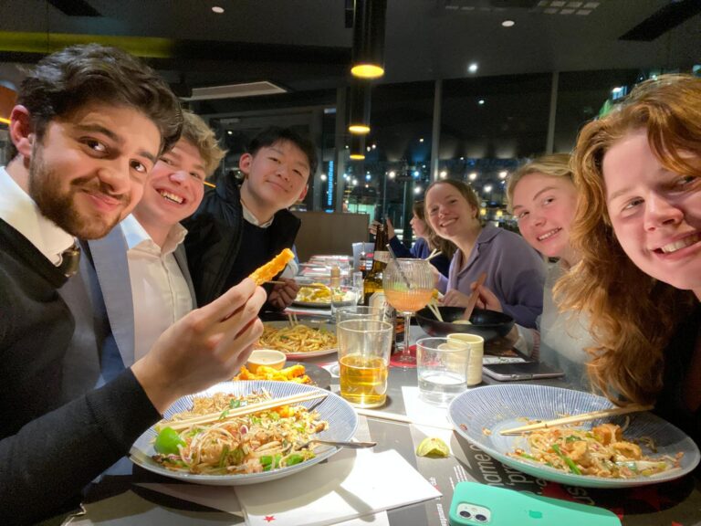Group of seven young adults laughing at dinner table with Asian noodle dishes and drinks, city lights visible through windows behind them.