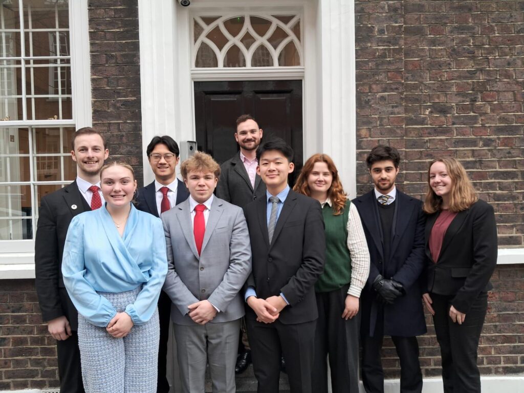 Nine young professionals in business clothing posed together outside a brick building entrance