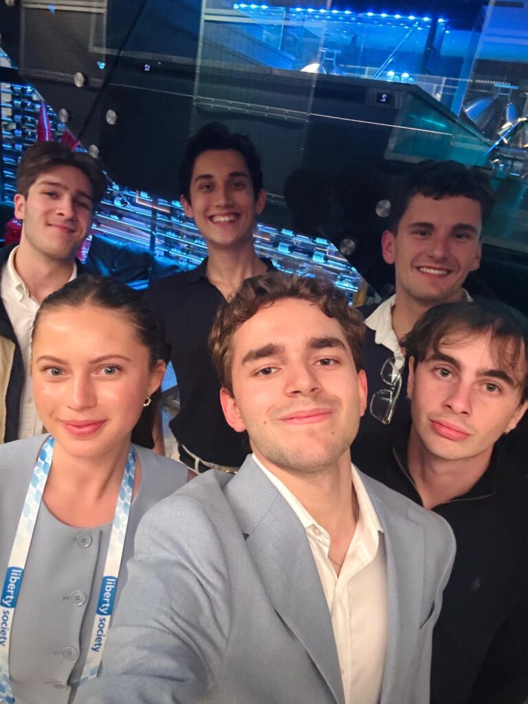 Six young adults posing together at a professional event venue with blue lighting in background