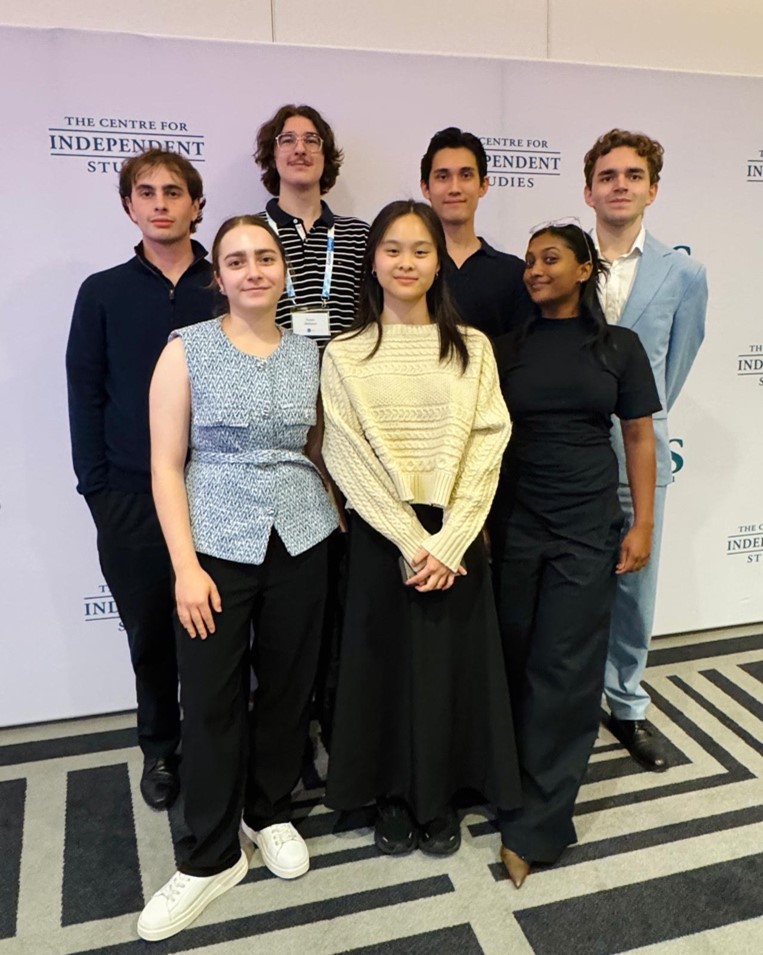 Seven young adults standing together in a group pose in front of The Centre for Independent Studies backdrop