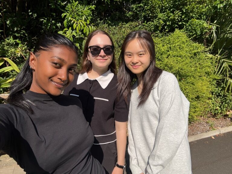 Three young women smiling together outdoors in front of green foliage
