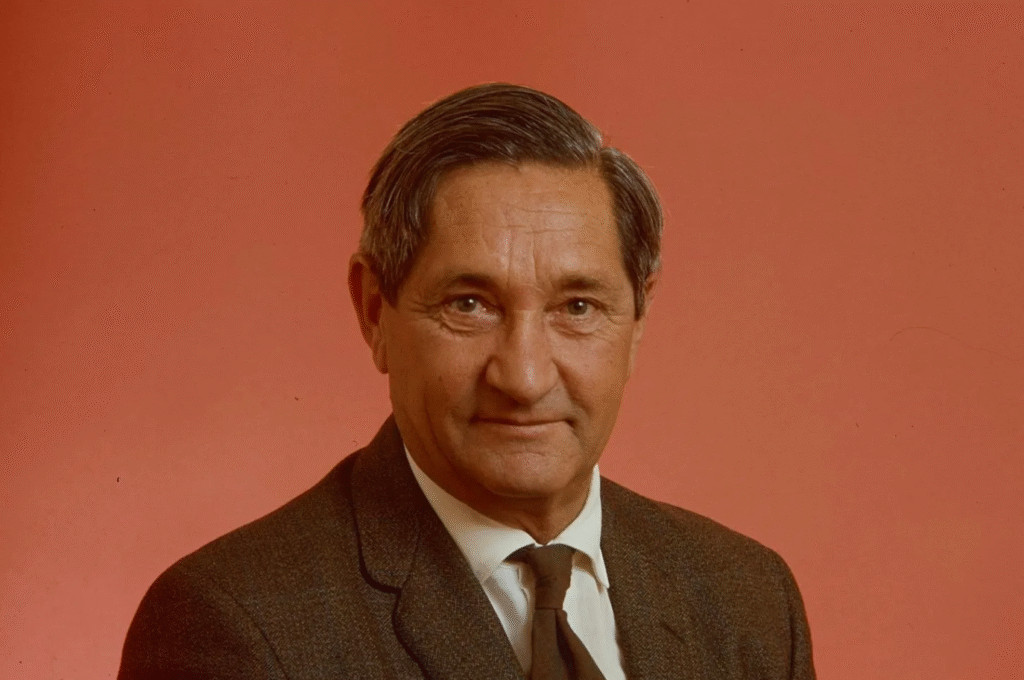 Man in brown suit and tie posed against coral-red studio background