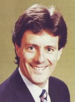 Professional headshot of man in suit and tie smiling at camera against beige background