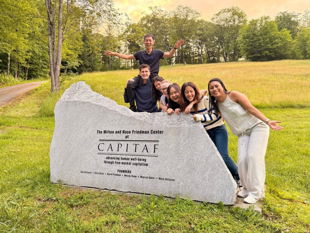 Six young adults posing around a stone monument marker for CAPITAF in a green field