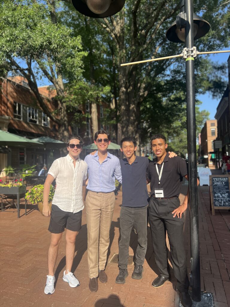 Four smiling men posing together on a brick plaza with historic buildings and trees in background