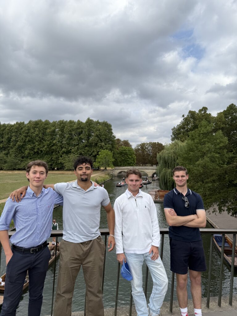 Four young men standing on a bridge over River Cam in Cambridge with trees and cloudy sky