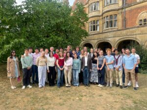 Large group of young adults standing in organized rows before a historic brick building with arched windows and ivy.