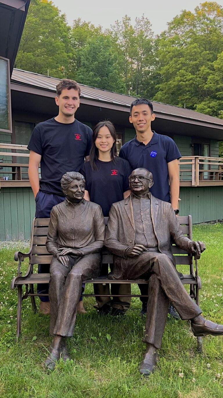 Three young adults standing behind bronze statues of two seated figures on a bench outdoors