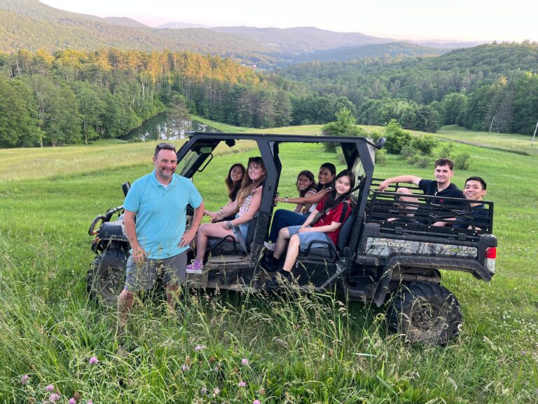 Group of 8 people smiling on a John Deere Gator in a grassy mountain valley with forested hills behind them
