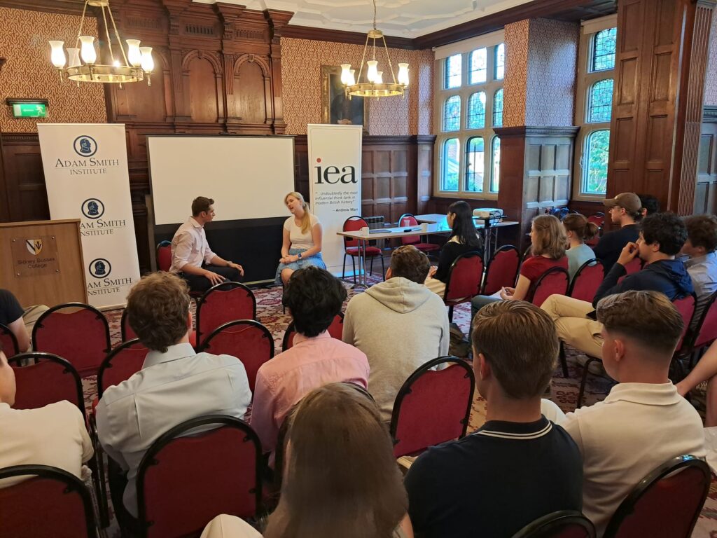 Panel discussion event with two speakers conversing in front of seated audience members in an elegant institutional setting.