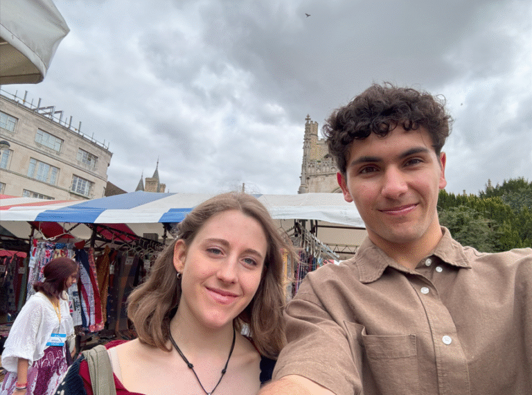 Two young adults smiling at outdoor market with historic buildings and market stalls in background